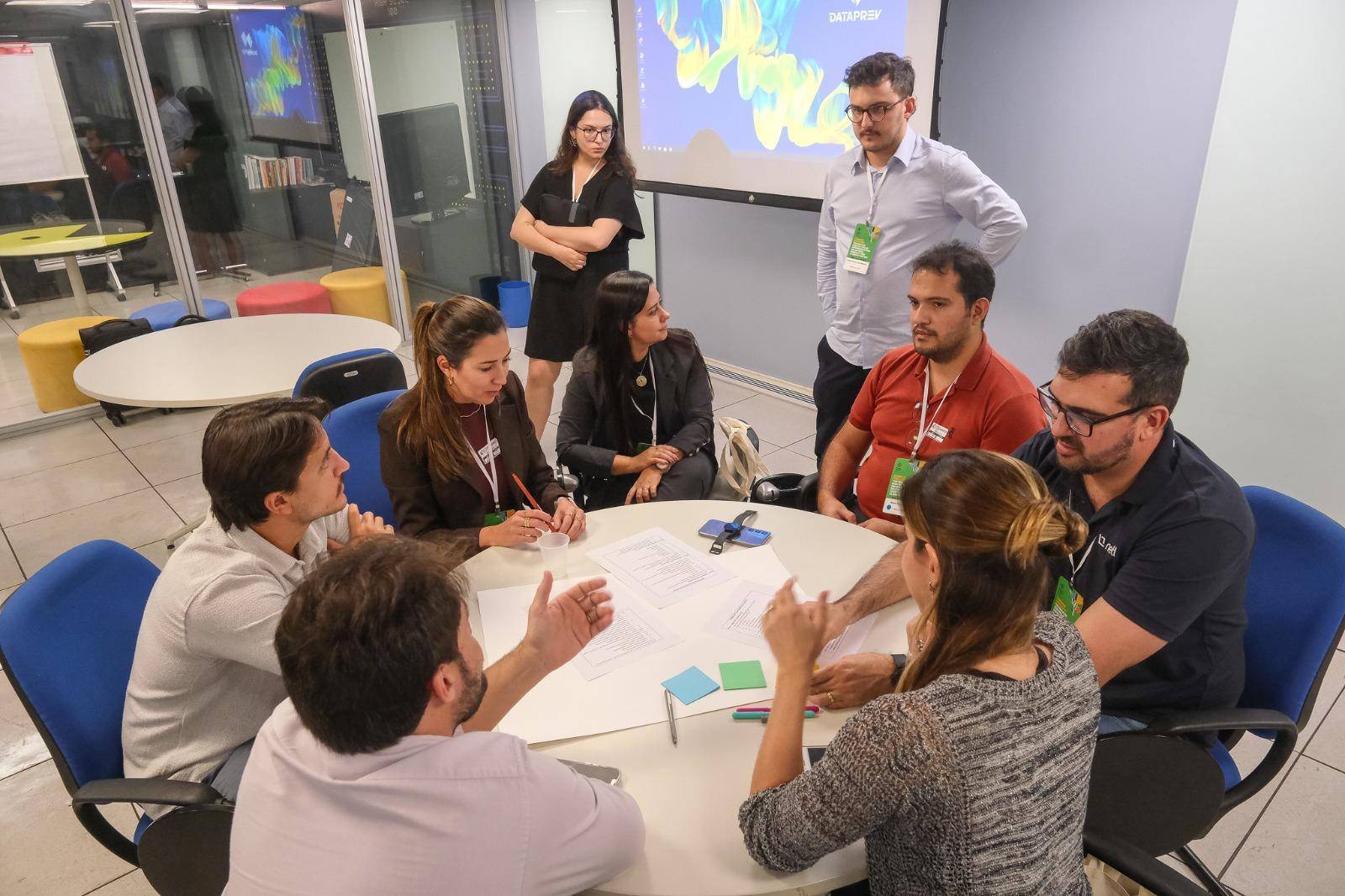 Nessa imagem podemos ver o grupo de trabalho ao redor de uma mesa. Existem sete  pessoas sentadas ao redor da mesa, enquanto outras duas observam o grupo de pé.